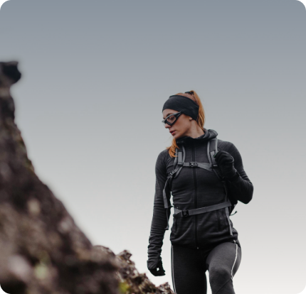 A woman in athletic gear and sunglasses hikes outdoors, wearing a backpack and looking to the side against a gray sky.