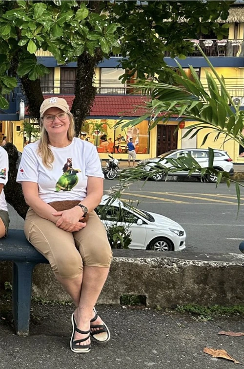 Woman wearing a white t-shirt and beige pants sits on a bench outdoors near a street, with trees and buildings in the background.