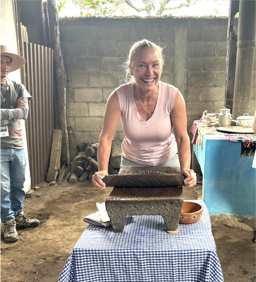A woman smiles while using a traditional stone grinder on a table covered with a checkered cloth, with two people and kitchen items in the background.