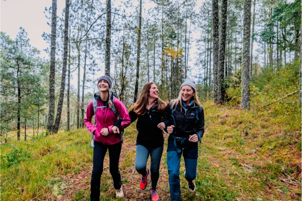 Three women wearing outdoor clothing hike and laugh together on a forest trail surrounded by tall trees and green grass.