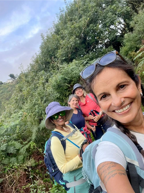 Four women in outdoor clothing pose for a selfie while hiking on a green, leafy trail with trees and shrubs in the background.