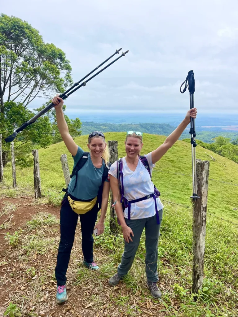 Two women in hiking gear stand on a grassy hilltop, smiling and raising trekking poles, with trees and a distant landscape in the background.