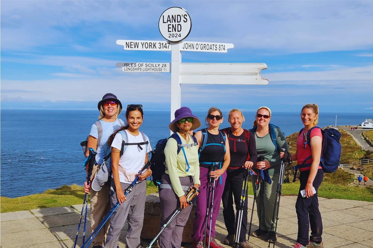 Seven women in hiking gear stand in front of the Land's End signpost with the ocean in the background on a clear day.