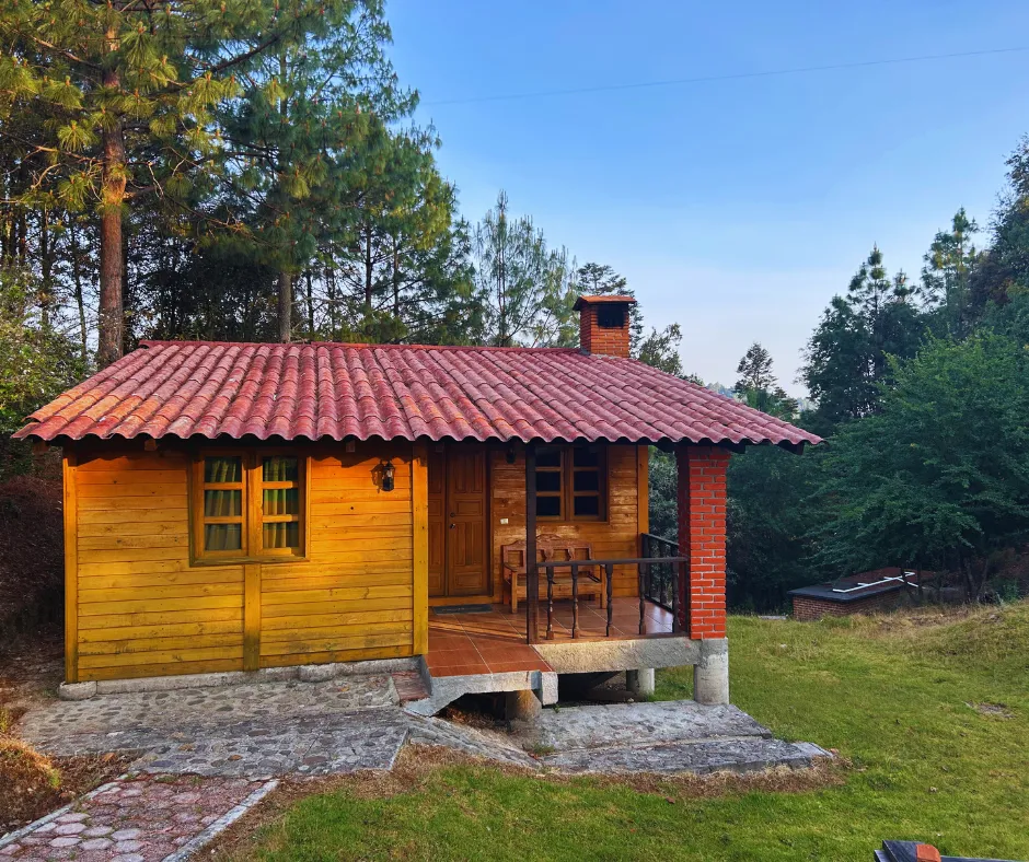 A small wooden cabin with a red tiled roof and front porch sits on a grassy area surrounded by trees under a clear sky.