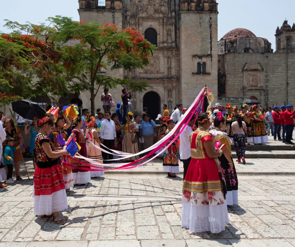 People in traditional Mexican attire perform a ribbon dance outdoors in front of a historic stone building, watched by a crowd.
