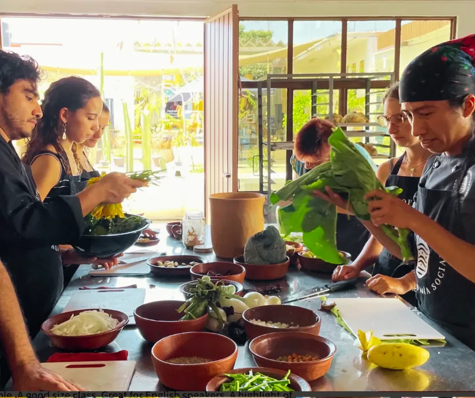 A group of people in a kitchen prepare food together at a table, surrounded by bowls of fresh vegetables and ingredients.