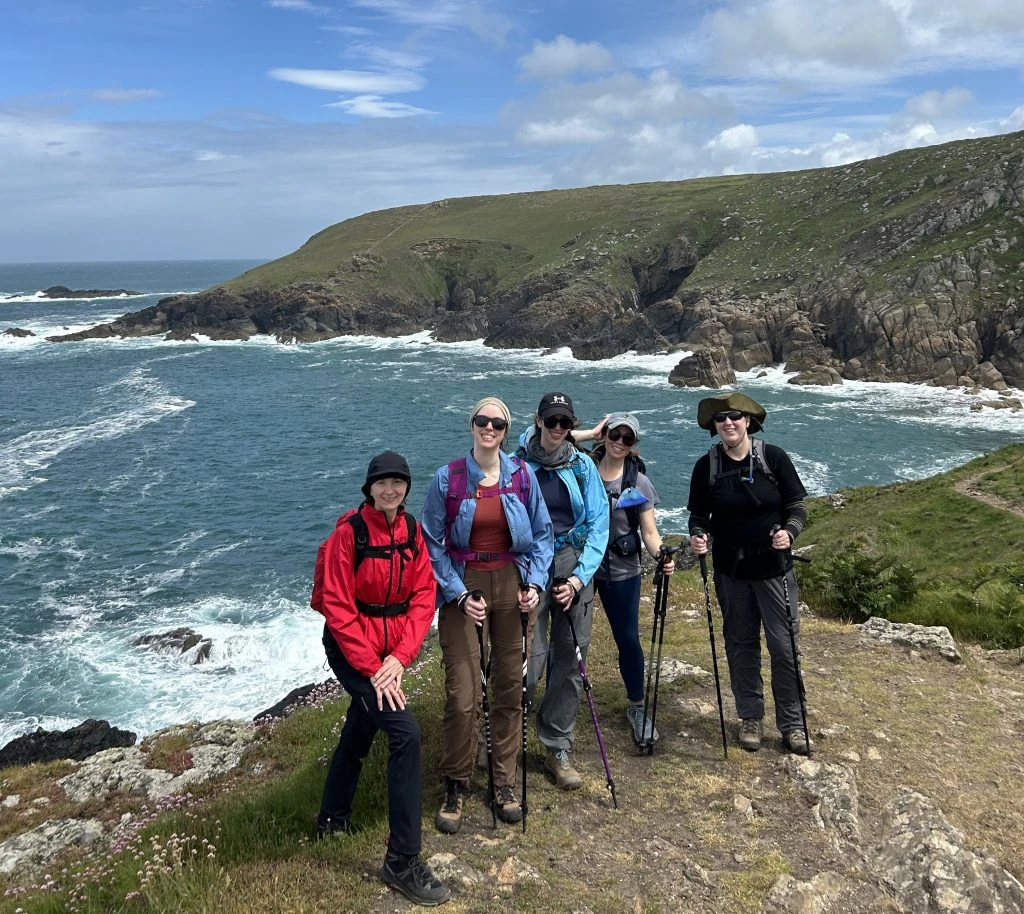 Five people with hiking gear stand on a grassy cliff overlooking the ocean with rocky hills and blue sky in the background.