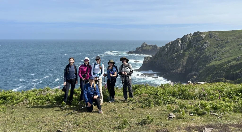 Six people in hiking gear stand and kneel on a grassy cliff overlooking a rocky coastline and the ocean under a partly cloudy sky.
