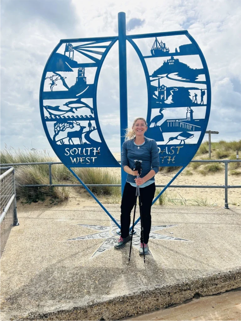 A woman stands smiling with walking poles in front of a blue metal sign shaped like a sail, labeled "SOUTH WEST COAST PATH," near a sandy beach with grass and cloudy sky.