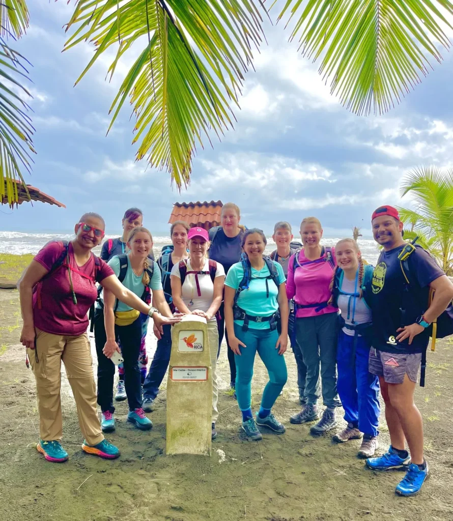 A group of ten people in hiking gear stand outdoors around a small concrete marker, with palm trees and the ocean in the background.
