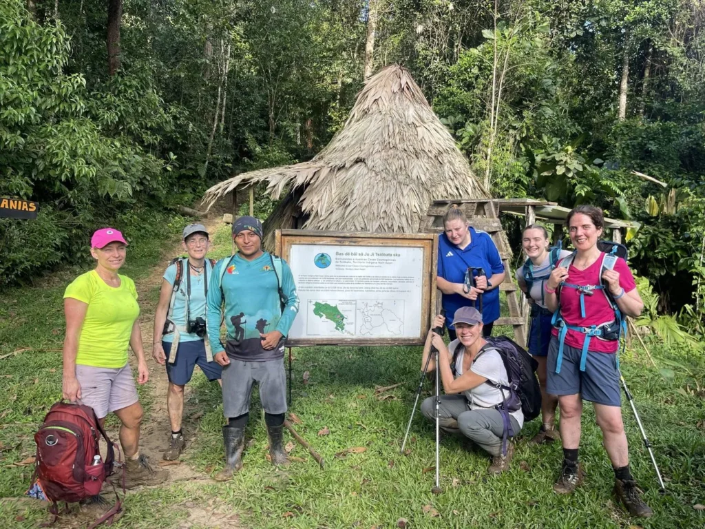 A group of seven hikers pose in front of an informational sign and a small thatched hut surrounded by dense greenery.