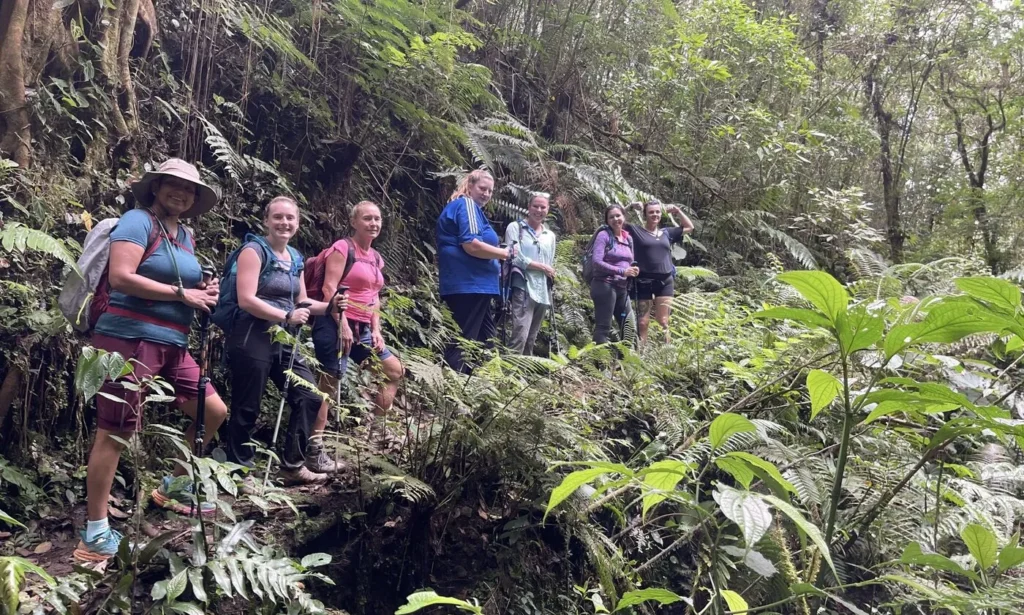 A group of seven hikers stand on a narrow, muddy trail surrounded by dense, green vegetation in a forest.