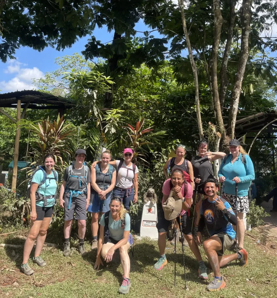 A group of ten hikers pose together outdoors in front of a lush, leafy background under sunny skies.