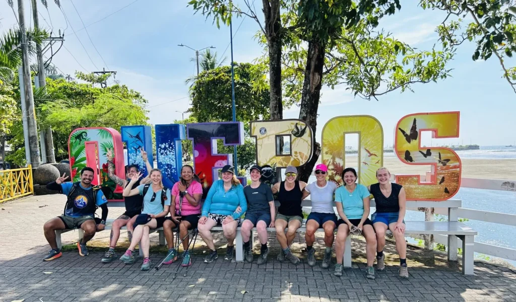 A group of twelve people sit and pose for a photo in front of large colorful &ldquo;QUEPOS&rdquo; letters on a sunny day, with trees and a waterfront visible in the background.