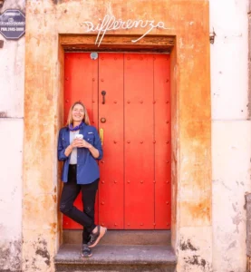 A person stands smiling and holding a cup in front of a large, bright red door set in a building with “Differenza” written above the doorway.