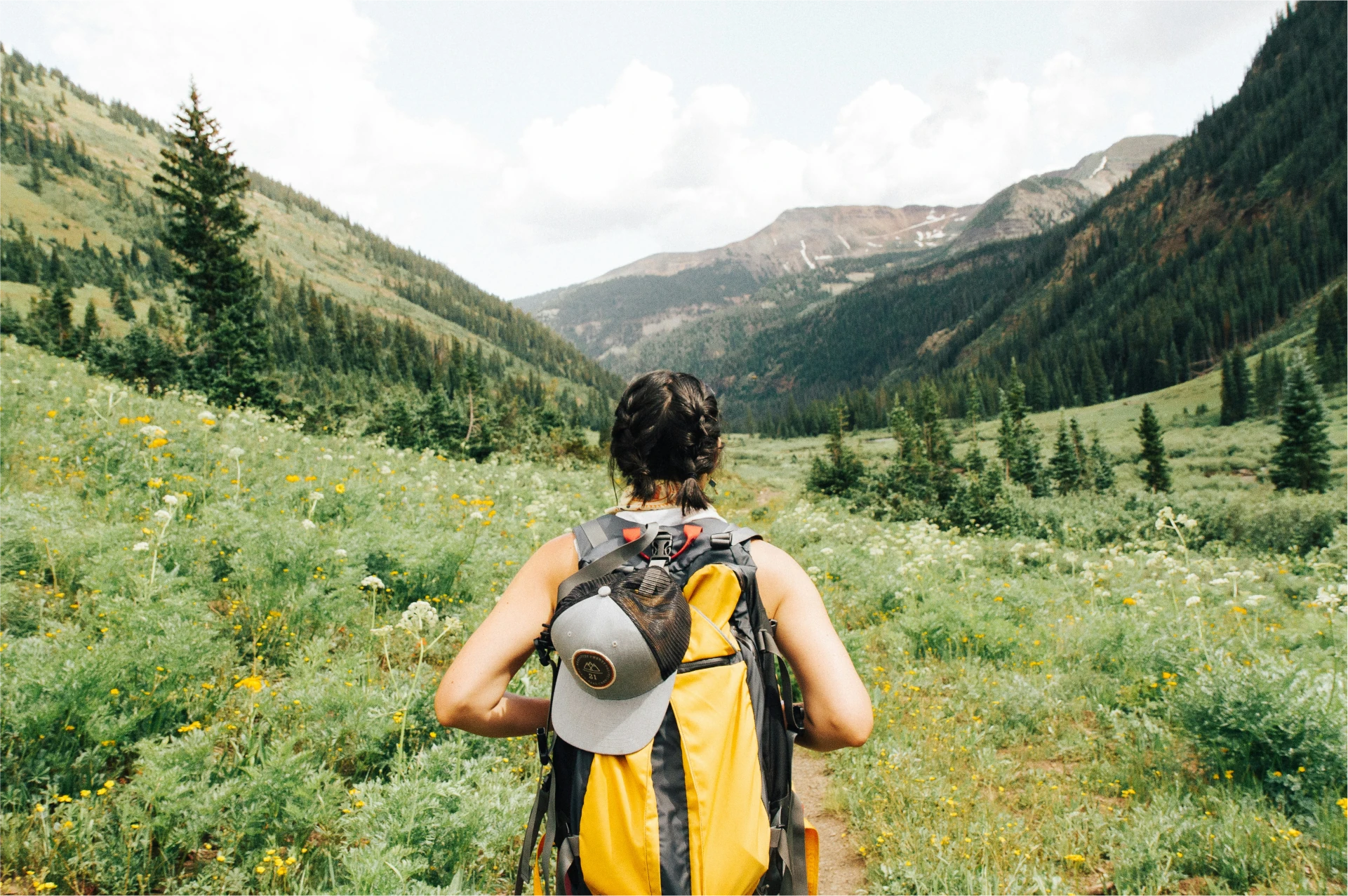 Person with a backpack and cap walks through a green mountain valley surrounded by trees and wildflowers under a partly cloudy sky.