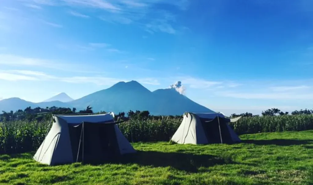 Two tents set up on grassy ground with mountains and a plume of smoke rising from one peak under a clear blue sky.