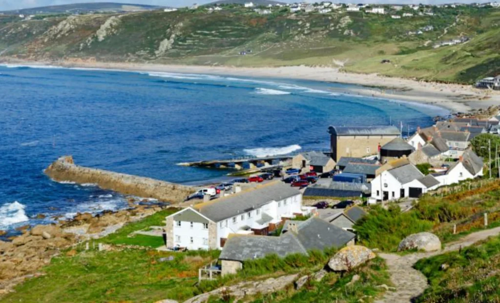 A coastal village with white houses sits beside a curved sandy beach and blue sea, with hills and scattered homes in the background.