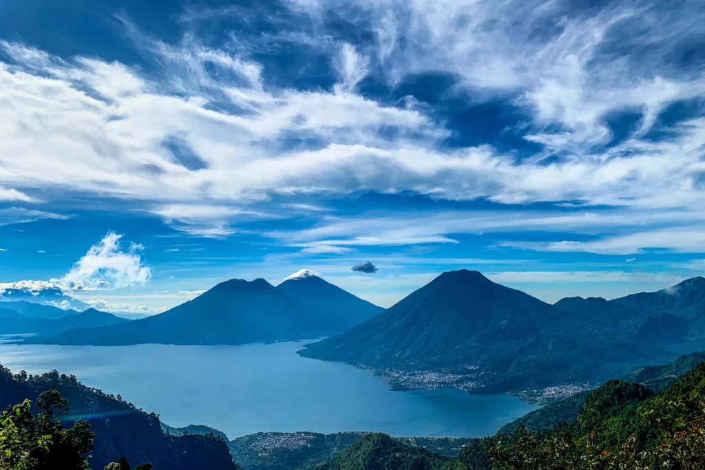 A scenic view of a lake surrounded by mountains and hills under a partly cloudy blue sky.