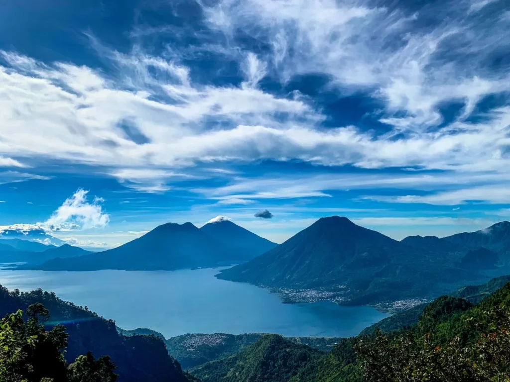 A wide lake is surrounded by green hills and volcanic mountains under a blue sky with scattered clouds.