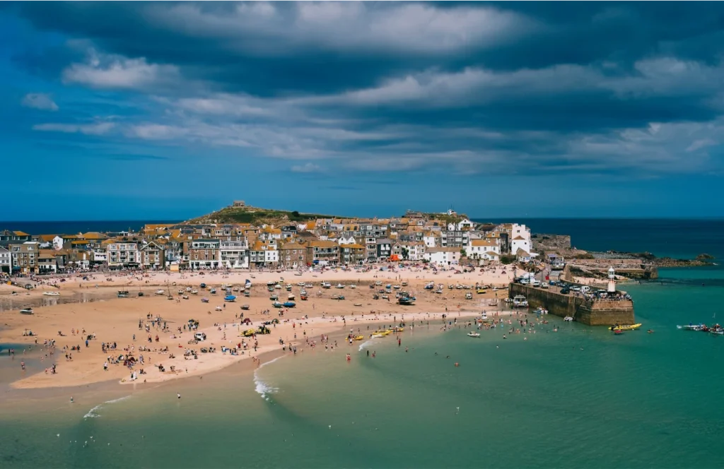 A coastal town with sandy beaches, people on the shore, historic buildings, and a cloudy sky above turquoise water.