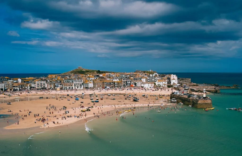 A coastal town with sandy beaches, clear turquoise water, and many people enjoying the shoreline under a cloudy sky. Buildings and a harbor are visible in the background.