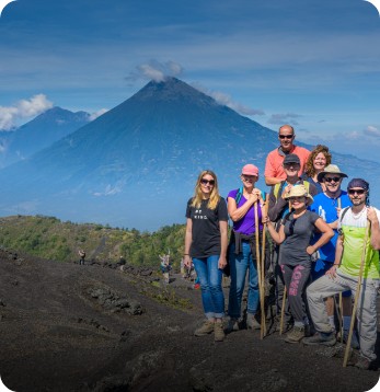 A group of hikers poses on a rocky slope with walking sticks, with a large volcano and blue sky in the background.