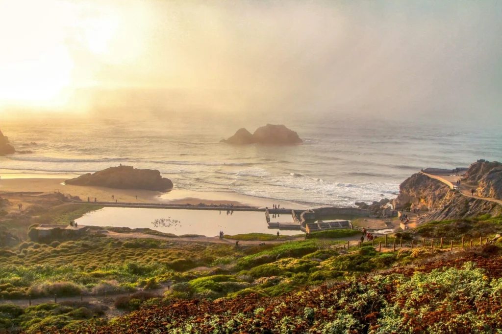 View of a rocky coastline with a rectangular tidal pool, people walking along paths, green vegetation, and ocean waves under a hazy sky.